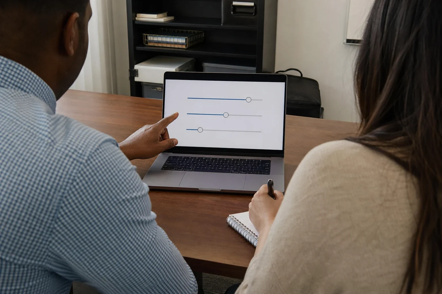 Over-the-shoulder view of a man pointing at slider options on a laptop while a woman takes notes in a spiral notebook at a wooden office desk